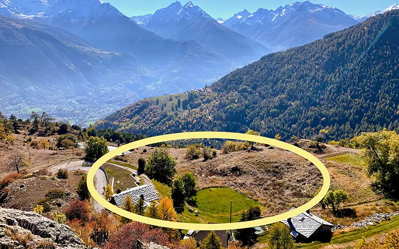 Een bergachtig landschap met groene velden, hier en daar wat bomen en huizen op de voorgrond. Een grote gele ovaal markeert een stuk open terrein vlakbij de huizen, met op de achtergrond de bergen.